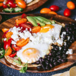 Huevos Rancheros made of sunny side up eggs, black beans, pico de gallo, cilantro a, and avocado on a wooden plate surrounded by cherry tomatoes and corn tortillas