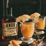 Glass mug of ho apple cider garnished with apple slices on the rim the mug resting on with slate coasters surrounded by freshly cut apples, loose Mulling spices in a silver scoop and a bottle of Knob Creek Whisky