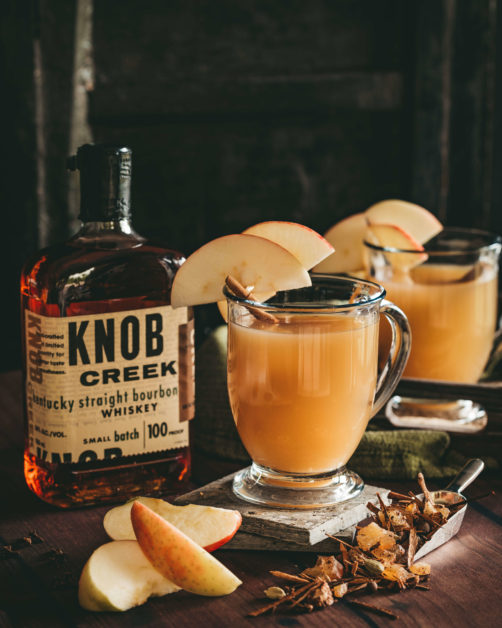 Glass mug of ho apple cider garnished with apple slices on the rim the mug resting on with slate coasters surrounded by freshly cut apples, loose Mulling spices in a silver scoop and a bottle of Knob Creek Whisky