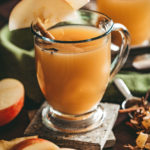 Glass mug of ho apple cider garnished with apple slices on the rim the mug resting on with slate coasters surrounded by freshly cut apples, loose Mulling spices in a silver scoop