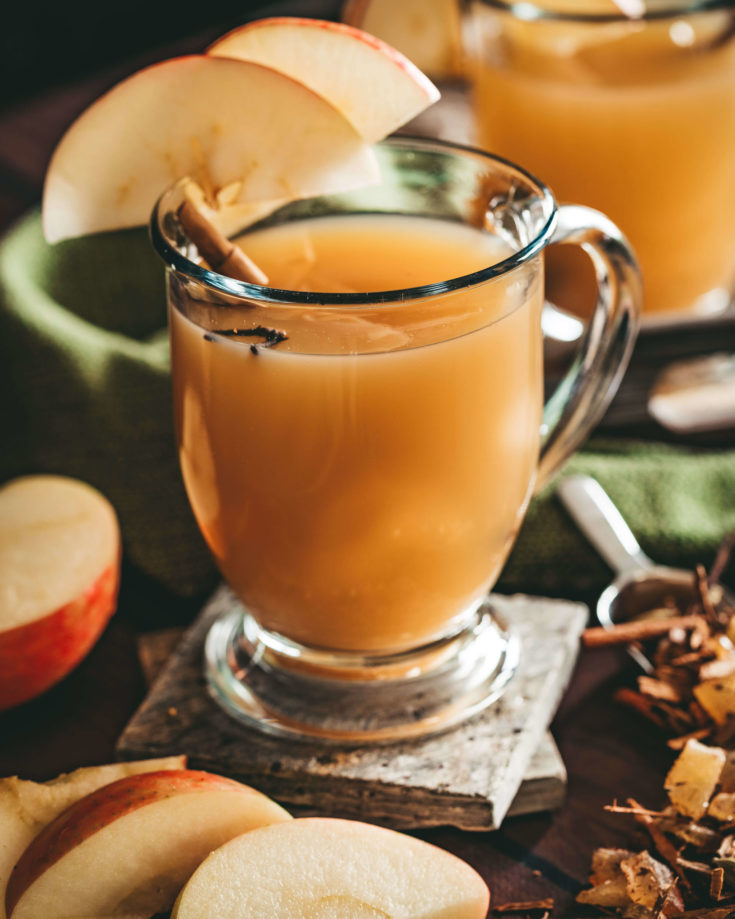 Glass mug of ho apple cider garnished with apple slices on the rim the mug resting on with slate coasters surrounded by freshly cut apples, loose Mulling spices in a silver scoop