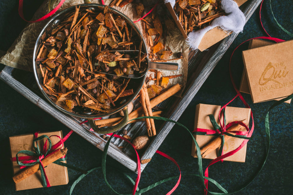Bowl filled with mixed mulling Spices on a tray surrounded by whole spices cinnamon, nutmeg, and allspice berries surrounded by red and green ribbon and small craft boxes wrapped like presents.