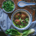 A blue and white Chinese porcelain bowl filled with Asian Meatball and Lemongrass Soup by The Qui Ingredient. The bowl is surrounded by a small circular plate of chopped cilantro some fresh bokchoy and a ladle soup spoon