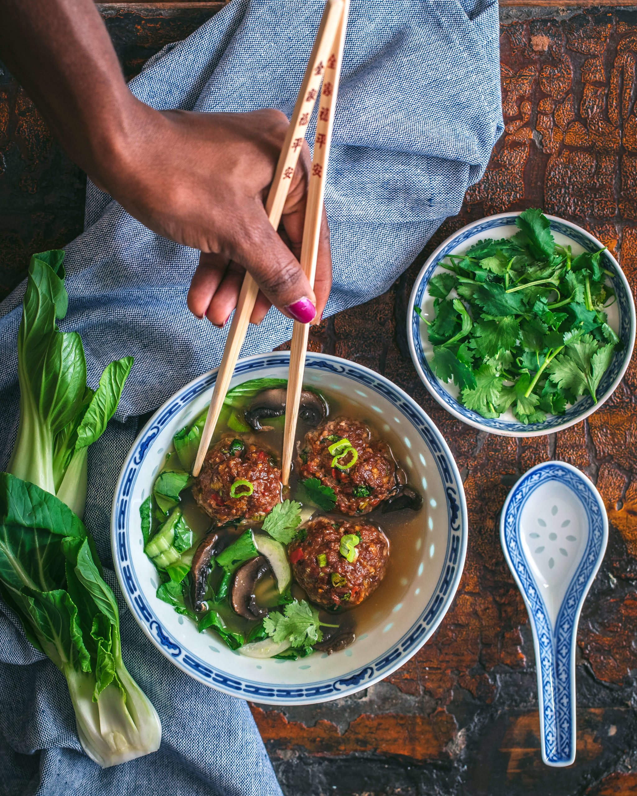 Hand holding wooden chopsticks over Chinese porcelain bowl filled with Asian Meatball soup by the Qui Ingredient surrounded by a small bowl of chopped cilantro, fresh bok choy and a ladle soup spoon.