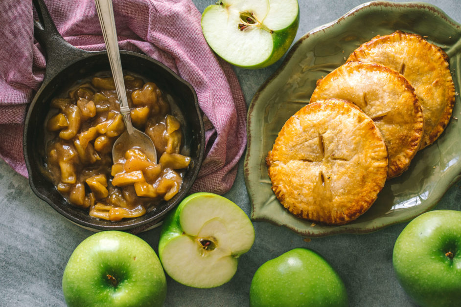 Horizontal image of Black lodge skillet with cooked apple pie filling wrapped in in a light red dish towel and next to it is a platter of apple hand pies surrounded by whole and sliced green apples on a marble slab table.