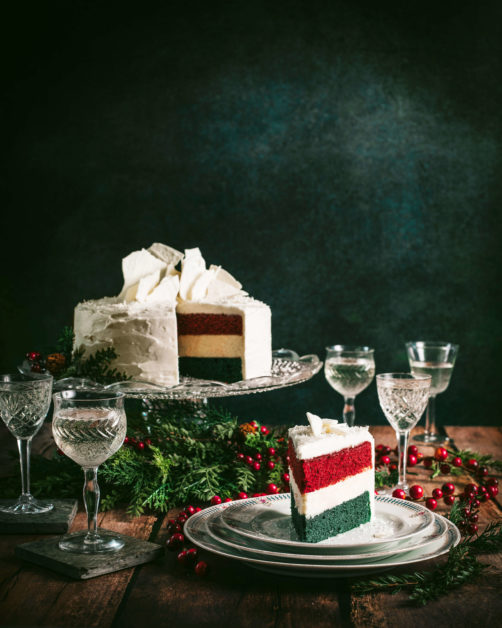 Three layer Red and Green Velvet Cheesecake with cream cheese icing on an crystal cake stand ganished red berries and mince sprigs surrounded by crystal coupe glasses filled with champagne