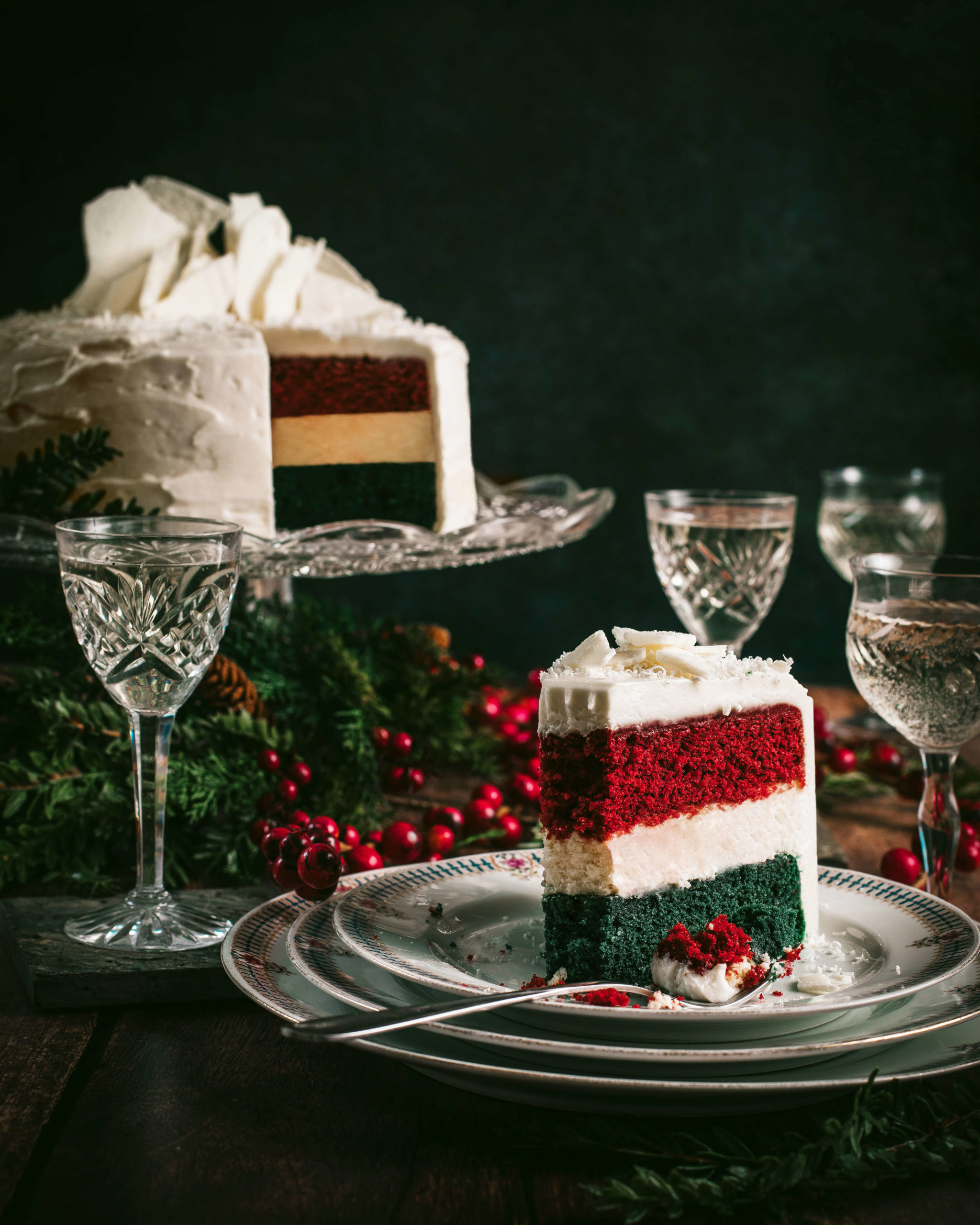Three layer Red and Green Velvet Cheesecake with cream cheese icing on an crystal cake stand ganished red berries and mince sprigs surrounded by crystal coupe glasses filled with champagne