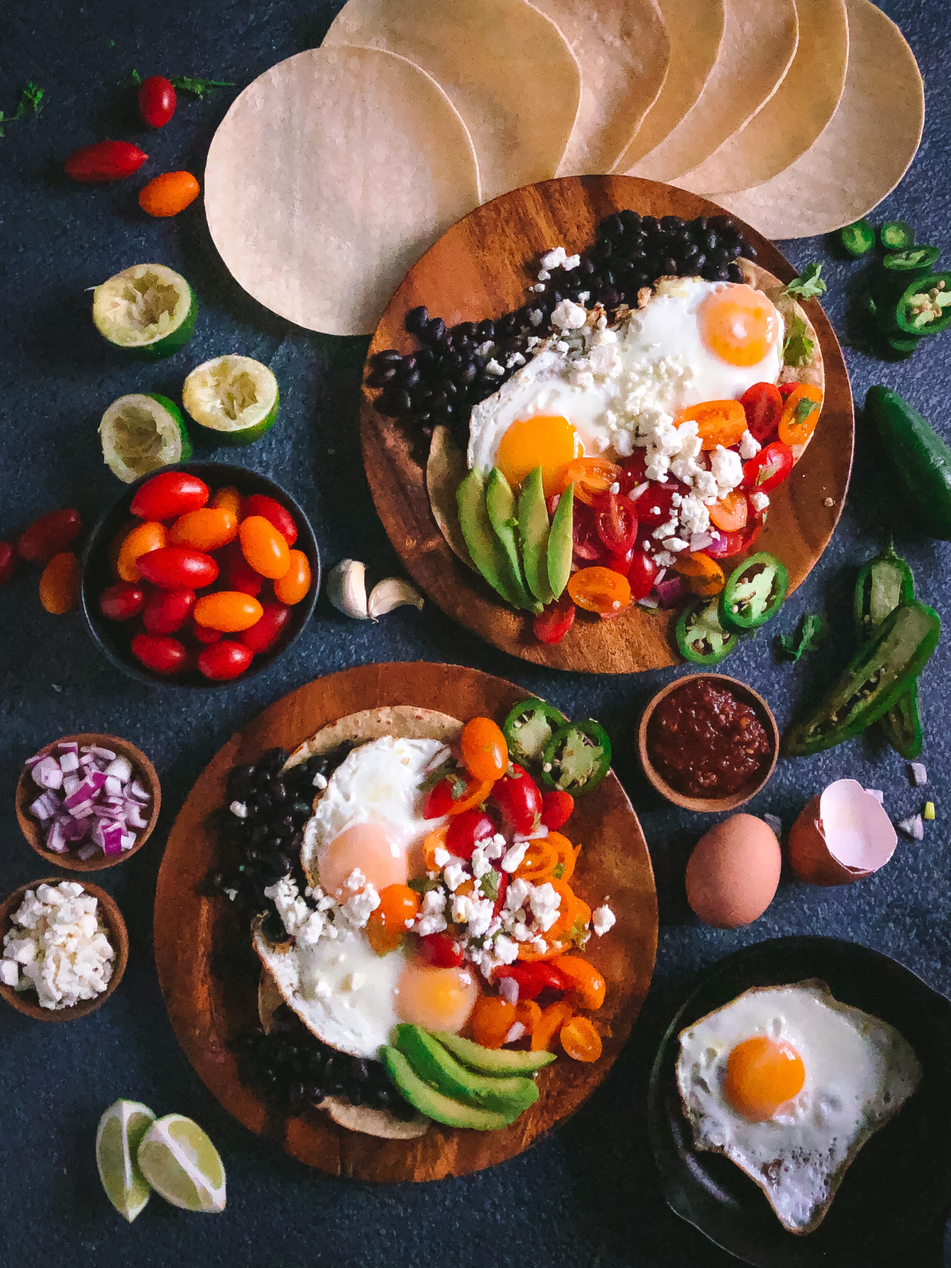 Huevos Rancheros made of sunny side up eggs, black beans, pico de gallo, cilantro a, and avocado on a wooden plate surrounded by cherry tomatoes and corn tortillas